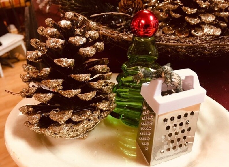 May include: A gold pine cone with glitter, a green glass bottle with a red top, and a small silver grater with a white handle.