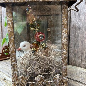 Puede incluir: Una linterna decorativa de estilo antiguo con un acabado dorado envejecido. En el interior, un p&aacute;jaro de cer&aacute;mica blanca se encuentra en un nido de musgo seco. Una hebra de cuentas con acentos naranjas y rojos cuelga de la parte superior.