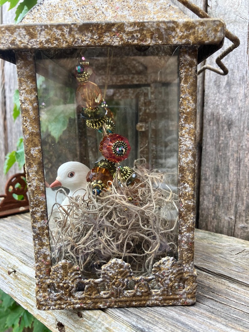 Puede incluir: Una linterna decorativa de estilo antiguo con un acabado dorado envejecido. En el interior, un p&aacute;jaro de cer&aacute;mica blanca se encuentra en un nido de musgo seco. Una hebra de cuentas con acentos naranjas y rojos cuelga de la parte superior.