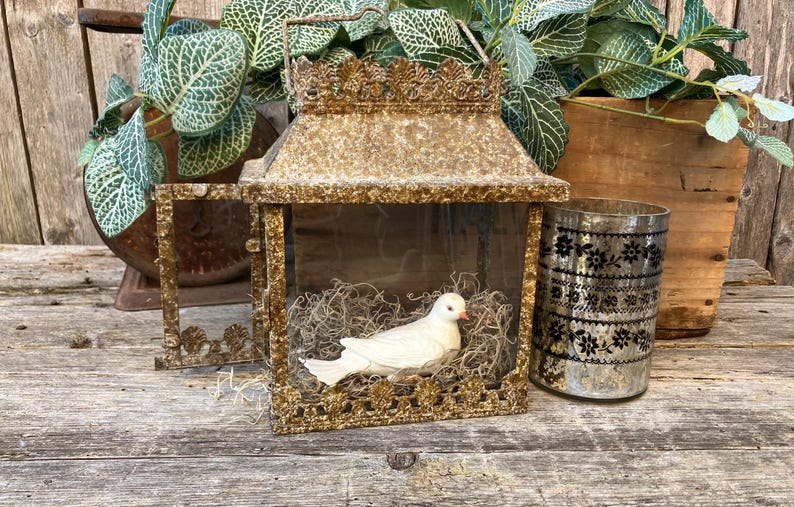 Puede incluir: Una linterna dorada de estilo vintage con un p&aacute;jaro decorativo en su interior, colocada sobre una superficie de madera envejecida. La linterna tiene una puerta con bisagras y est&aacute; junto a un jarr&oacute;n de cristal decorativo y plantas en maceta.