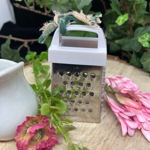 May include: A small, white and silver grater with a handle. The grater has a green and white fabric tied around the handle.