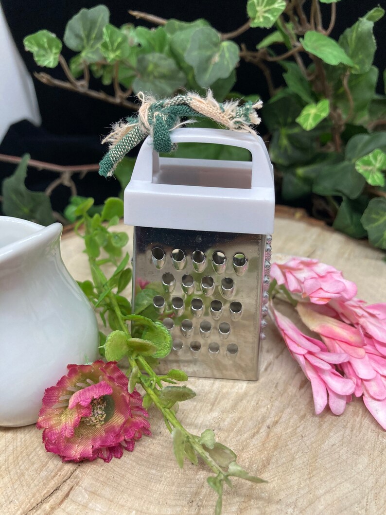 May include: A small, white and silver grater with a handle. The grater has a green and white fabric tied around the handle.