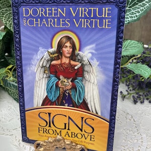 May include: A book titled "Signs From Above" by Doreen Virtue and Charles Virtue. The cover features an illustration of a woman with angel wings, a halo, and butterflies. The book is framed in a blue border. Two gold-colored decorative objects are in the foreground.