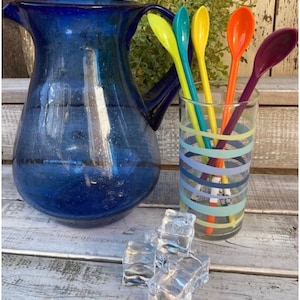 May include: A blue glass pitcher and a striped glass with colorful spoons. The spoons are yellow, teal, orange, and purple. Clear ice cubes are in front of the pitcher and glass. The pitcher has a handle and a wide spout.