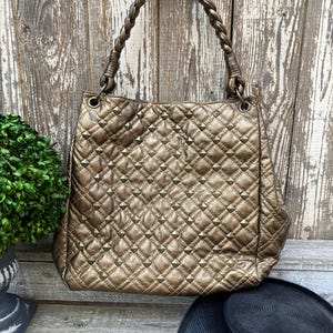 May include: A bronze-colored quilted handbag with pyramid-shaped studs. The bag has a braided handle and is set against a weathered wooden background. A black hat is visible in the foreground.