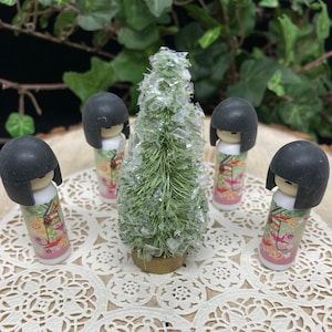 May include: Four small black-haired dolls wearing colorful kimonos are arranged around a miniature green Christmas tree with white glitter. The dolls are standing on a white lace doily.