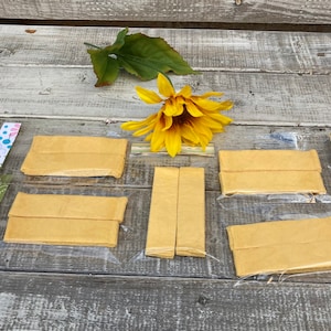 May include: Six packages of yellow paper strips, each package containing two strips, are arranged on a wooden surface. A single artificial sunflower is placed above the packages.
