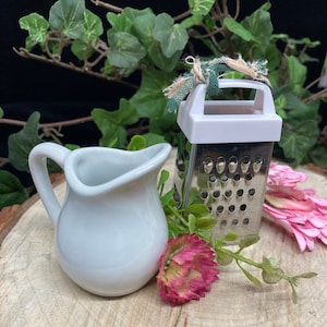 May include: A white ceramic creamer with a handle sits next to a small silver grater with a white plastic top. The grater has a green and white fabric tied around the handle. The objects are on a wooden surface with green leaves and pink flowers.