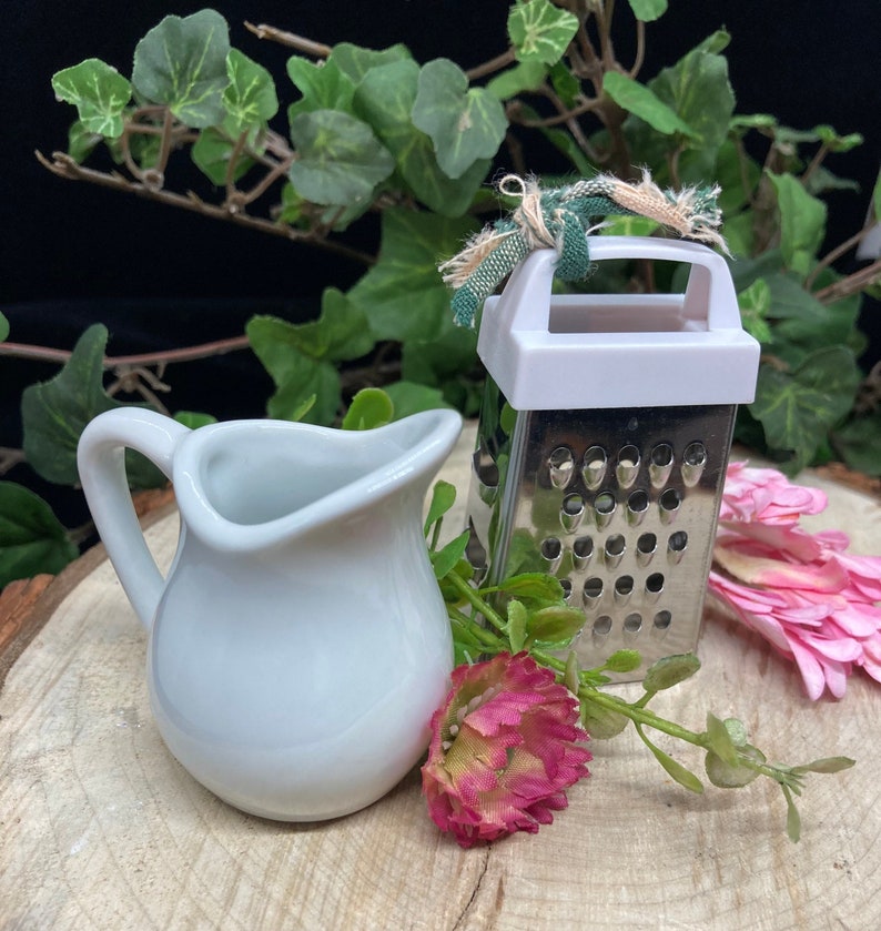 May include: A white ceramic creamer with a handle sits next to a small silver grater with a white plastic top. The grater has a green and white fabric tied around the handle. The objects are on a wooden surface with green leaves and pink flowers.