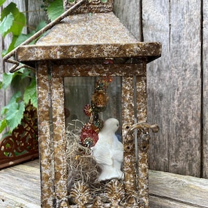 Puede incluir: Una linterna de metal de estilo vintage con un acabado dorado envejecido. En el interior, una figura de paloma de cer&aacute;mica blanca descansa entre musgo seco y cuentas decorativas. La linterna tiene una puerta con bisagras y un asa.
