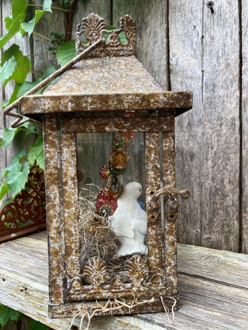 Puede incluir: Una linterna de metal de estilo vintage con un acabado dorado envejecido. En el interior, una figura de paloma de cer&aacute;mica blanca descansa entre musgo seco y cuentas decorativas. La linterna tiene una puerta con bisagras y un asa.