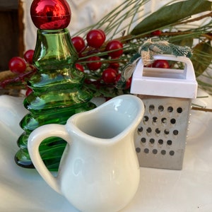 May include: A white ceramic creamer, a green glass Christmas tree bottle, and a silver grater with a white plastic handle. The items are arranged on a white surface with red berries and green pine branches.