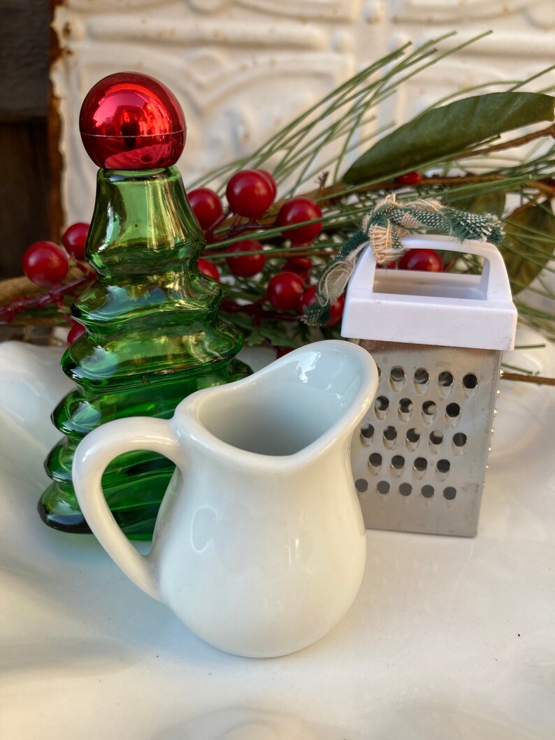 May include: A white ceramic creamer, a green glass Christmas tree bottle, and a silver grater with a white plastic handle. The items are arranged on a white surface with red berries and green pine branches.