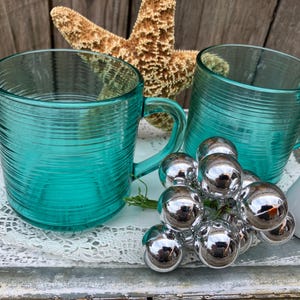 May include: Two teal blue glass mugs with a ribbed design. The mugs are sitting on a white lace doily on a wooden surface. There are silver ornaments in the center of the image.