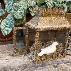 Puede incluir: Una linterna decorativa de estilo antiguo con una figura de paloma blanca en su interior. La linterna tiene un marco dorado y ornamentado y se encuentra sobre una superficie de madera envejecida. Tambi&eacute;n hay una balanza de estilo vintage y un vaso con un dise&ntilde;o floral negro.