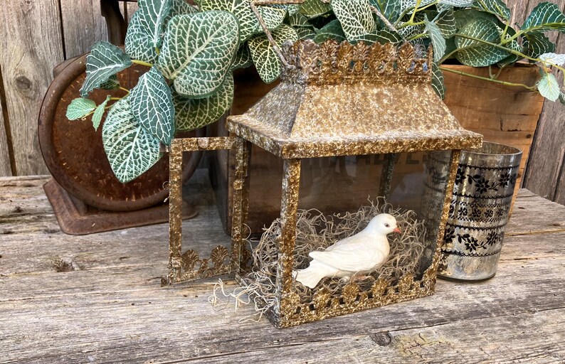 Puede incluir: Una linterna decorativa de estilo antiguo con una figura de paloma blanca en su interior. La linterna tiene un marco dorado y ornamentado y se encuentra sobre una superficie de madera envejecida. Tambi&eacute;n hay una balanza de estilo vintage y un vaso con un dise&ntilde;o floral negro.