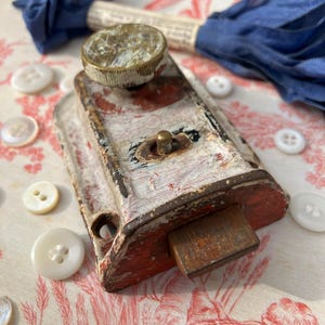 May include: An antique, distressed metal latch with red and white paint. A small, round brass piece sits on top. White buttons and a blue ribbon are scattered around the latch on a patterned fabric.