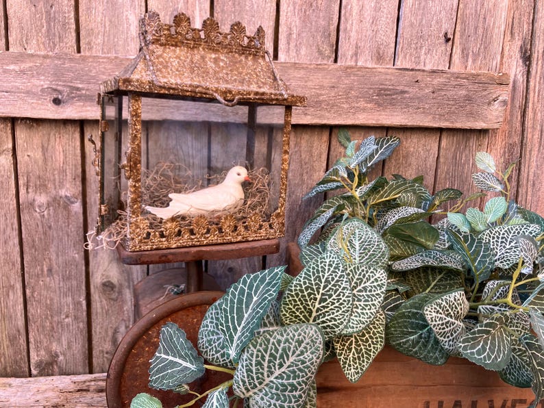 Puede incluir: Una jaula decorativa con una figura de paloma blanca descansando sobre un nido. La jaula ornamentada, de estilo antiguo, est&aacute; hecha de metal dorado y se encuentra sobre un fondo de madera r&uacute;stica. Plantas de hojas verdes y blancas en primer plano.