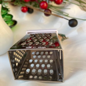 May include: A silver metal grater with a square shape and a small handle. The grater has a fine mesh pattern and is designed for grating cheese or other foods.