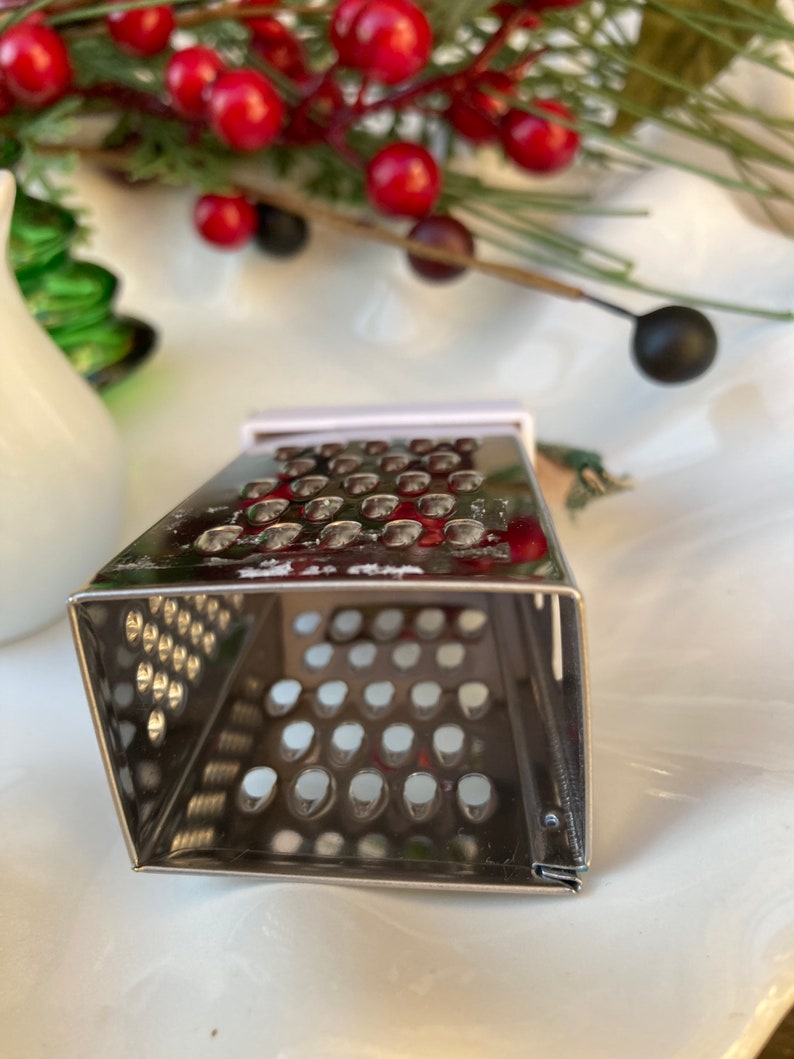 May include: A silver metal grater with a square shape and a small handle. The grater has a fine mesh pattern and is designed for grating cheese or other foods.