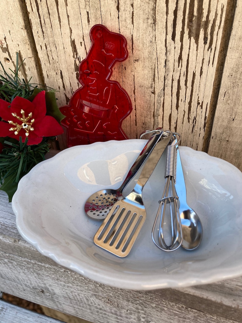 Op de afbeelding: Een set miniatuur keukengerei in een witte keramische kom. De keukengerei omvatten een spatel, een garde, een lepel en een schuimspaan. De keukengerei zijn gemaakt van zilverkleurig metaal en hebben een glanzende afwerking.