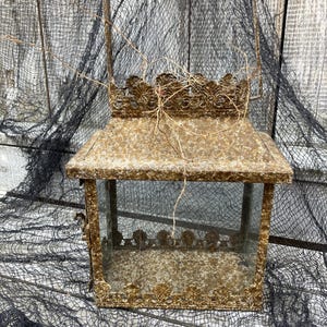 Puede incluir: Una linterna rectangular de estilo vintage con un dise&ntilde;o ornamentado. La linterna es de metal con un acabado dorado envejecido y paneles de vidrio transparente. Cuenta con un asa para transportar y es adecuada para velas o elementos decorativos.