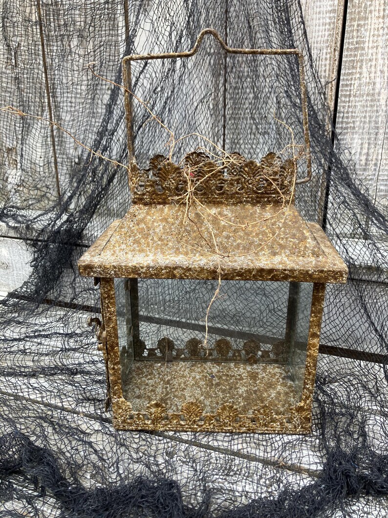 Puede incluir: Una linterna rectangular de estilo vintage con un dise&ntilde;o ornamentado. La linterna es de metal con un acabado dorado envejecido y paneles de vidrio transparente. Cuenta con un asa para transportar y es adecuada para velas o elementos decorativos.