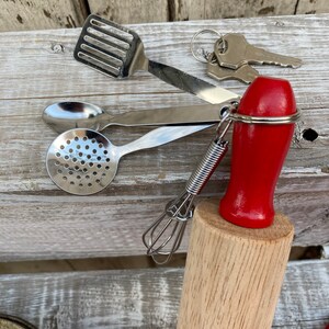 Op de afbeelding: Een set miniatuur keukengerei, waaronder een deegroller, een garde, een schuimspaan, een lepel en een spatel. Het keukengerei is bevestigd aan een sleutelhanger met een rode handgreep.