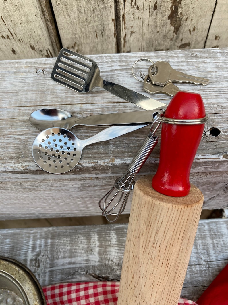 Op de afbeelding: Een set miniatuur keukengerei, waaronder een deegroller, een garde, een schuimspaan, een lepel en een spatel. Het keukengerei is bevestigd aan een sleutelhanger met een rode handgreep.