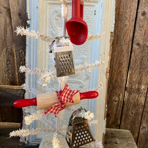 May include: A white artificial Christmas tree decorated with kitchen utensils, including a red scoop, a rolling pin, and two silver cheese graters. The tree is in a white pot and is sitting on a wooden surface.