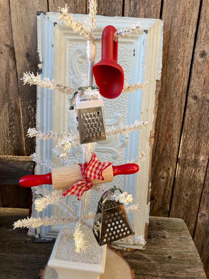 May include: A white artificial Christmas tree decorated with kitchen utensils, including a red scoop, a rolling pin, and two silver cheese graters. The tree is in a white pot and is sitting on a wooden surface.