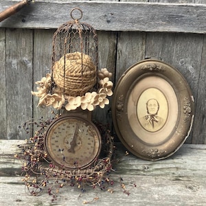 May include: An antique scale with a decorative birdcage and a framed portrait. The scale has a round face with numbers and a needle. The birdcage holds a woven sphere and is adorned with flowers. A portrait in an oval frame is next to the scale.