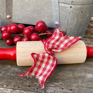 May include: A red and white checkered ribbon tied around a wooden rolling pin with red handles. The rolling pin is lying on a wooden surface.