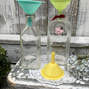 May include: Clear glass bottles with colorful funnels. One bottle has a teal funnel, another has a yellow-green funnel, and a third bottle has a small yellow funnel. The bottles are decorated with a pink flower and a red ribbon. The bottles are on a white lace doily.