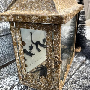 Puede incluir: Una linterna rectangular r&uacute;stica con un acabado dorado envejecido. La linterna presenta una puerta con bisagras y un panel de vidrio que muestra siluetas de gatos negros y el texto "happy halloween". Los elementos decorativos incluyen una ara&ntilde;a y una red negra.