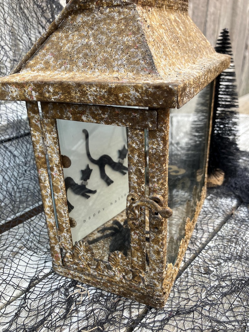 Puede incluir: Una linterna rectangular r&uacute;stica con un acabado dorado envejecido. La linterna presenta una puerta con bisagras y un panel de vidrio que muestra siluetas de gatos negros y el texto "happy halloween". Los elementos decorativos incluyen una ara&ntilde;a y una red negra.