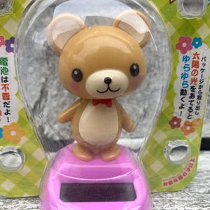 May include: A solar-powered bear figurine with a red bow tie, standing on a pink base. The bear is enclosed in a clear plastic bubble, with Japanese text on the packaging. The packaging has a green and white checkered pattern with floral accents.