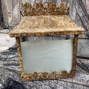 Puede incluir: Una linterna rectangular de estilo vintage con un acabado dorado envejecido. La linterna presenta adornos decorativos ornamentados a lo largo de los bordes superior e inferior. Los lados est&aacute;n abiertos, revelando un interior azul claro. Es un art&iacute;culo decorativo.