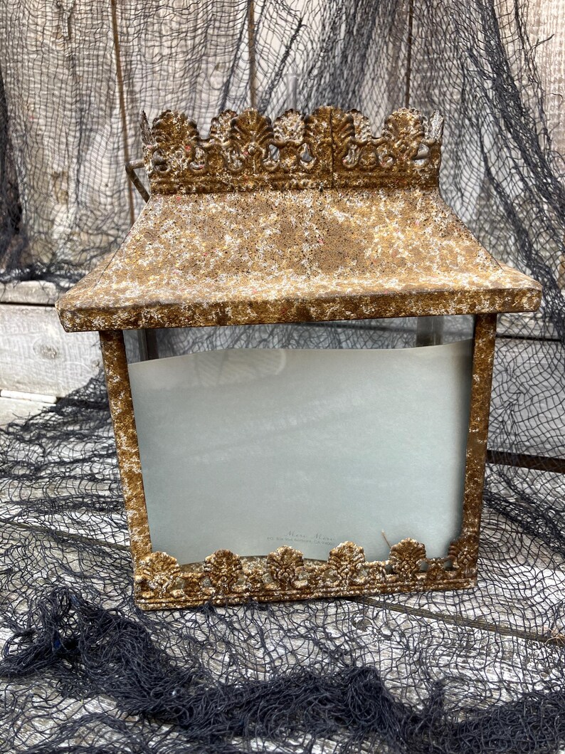 Puede incluir: Una linterna rectangular de estilo vintage con un acabado dorado envejecido. La linterna presenta adornos decorativos ornamentados a lo largo de los bordes superior e inferior. Los lados est&aacute;n abiertos, revelando un interior azul claro. Es un art&iacute;culo decorativo.