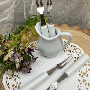 May include: Four white handled forks with silver tines. Two forks are in a white ceramic pitcher, and two forks are on a white doily.