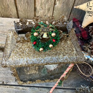 Puede incluir: Una caja rectangular dorada con tem&aacute;tica navide&ntilde;a con una corona verde decorativa, bayas rojas y adornos dorados. Un adorno en forma de estrella con el texto "Let it Snow" es visible.