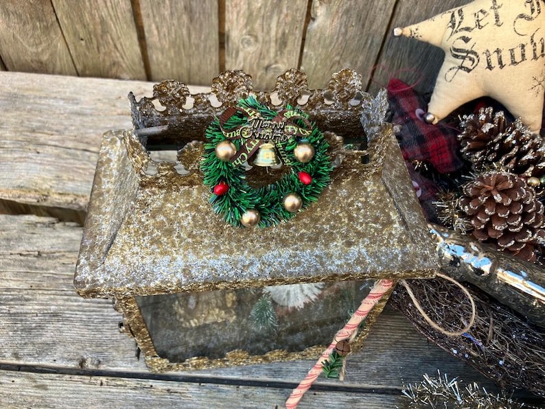 Puede incluir: Una caja rectangular dorada con tem&aacute;tica navide&ntilde;a con una corona verde decorativa, bayas rojas y adornos dorados. Un adorno en forma de estrella con el texto "Let it Snow" es visible.