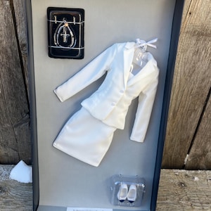 May include: A white doll outfit with a jacket, skirt, and shoes. The outfit is packaged in a black box with a clear plastic insert. The box has instructions on how to remove the outfit from the box.