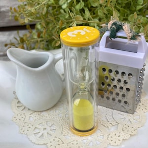 May include: A yellow 3-minute hourglass timer with a white and silver grater in the background.