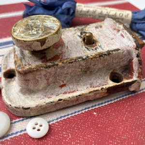 May include: An antique, distressed metal latch painted white with red peeking through. A brass knob is on top, and white buttons are scattered around. The latch is on a red and white striped surface.