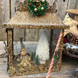 Puede incluir: Una linterna decorativa de estilo antiguo con un marco dorado. En el interior, se exhiben una figura de mu&ntilde;eco de nieve, &aacute;rboles de Navidad en miniatura y un &aacute;rbol de cepillo blanco. Un bast&oacute;n de caramelo y una peque&ntilde;a corona adornan la linterna.