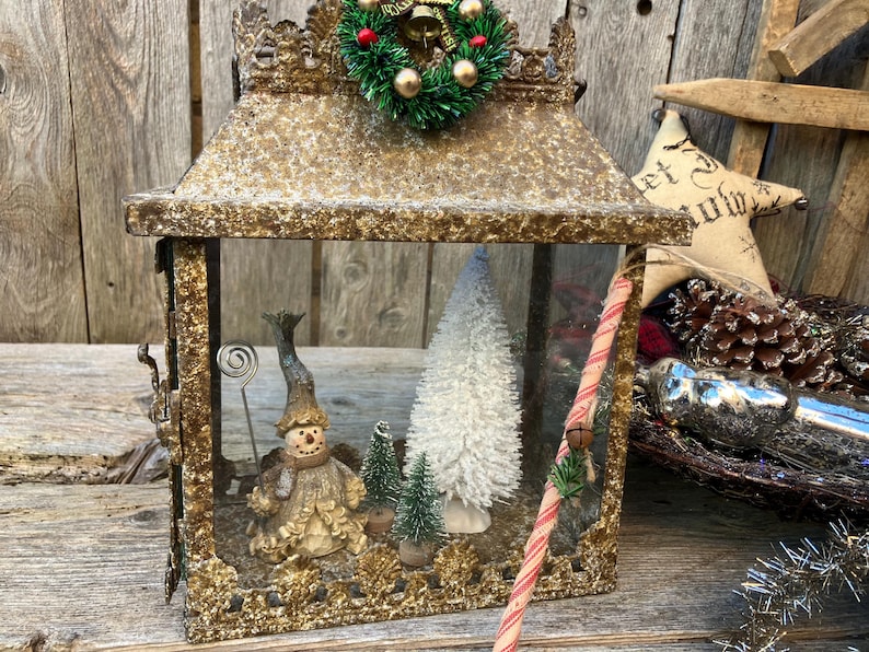 Puede incluir: Una linterna decorativa de estilo antiguo con un marco dorado. En el interior, se exhiben una figura de mu&ntilde;eco de nieve, &aacute;rboles de Navidad en miniatura y un &aacute;rbol de cepillo blanco. Un bast&oacute;n de caramelo y una peque&ntilde;a corona adornan la linterna.