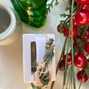 May include: White plastic bottle stopper with a green and brown rope threaded through it. The stopper is sitting on a white surface with a green glass bottle and red berries in the background.