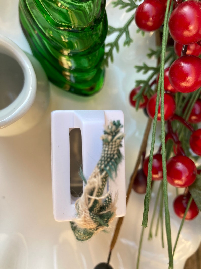 May include: White plastic bottle stopper with a green and brown rope threaded through it. The stopper is sitting on a white surface with a green glass bottle and red berries in the background.
