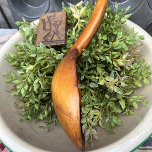 May include: A long-handled wooden ladle with a curved hook at the end of the handle. The ladle is placed inside a shallow, light-colored bowl filled with green foliage. A small, rectangular wooden block with a carved figure is also present.